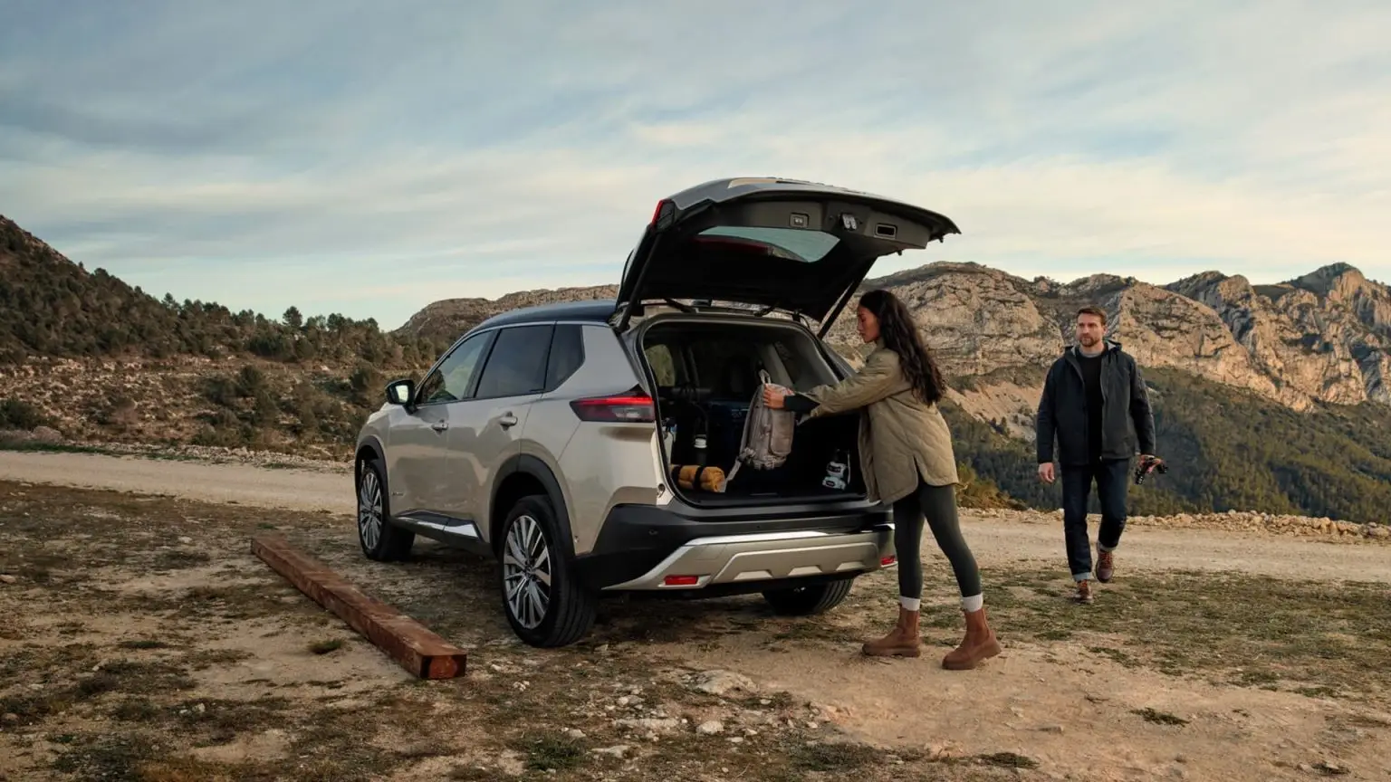 Ein Paar lädt Outdoor-Ausrüstung in den Kofferraum eines Nissan X-Trail vor Bergkulisse.