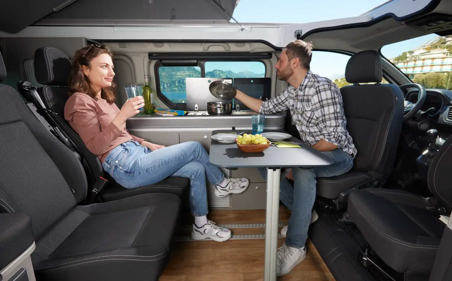 Paar sitzt im Wohnraum eines Nissan Primastar mit Esstisch, Kochnische und offener Seitentür.