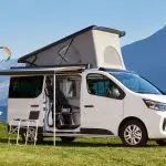 Weißer Nissan Primastar Campervan mit aufgeklapptem Aufstelldach und Campingmöbeln am See.