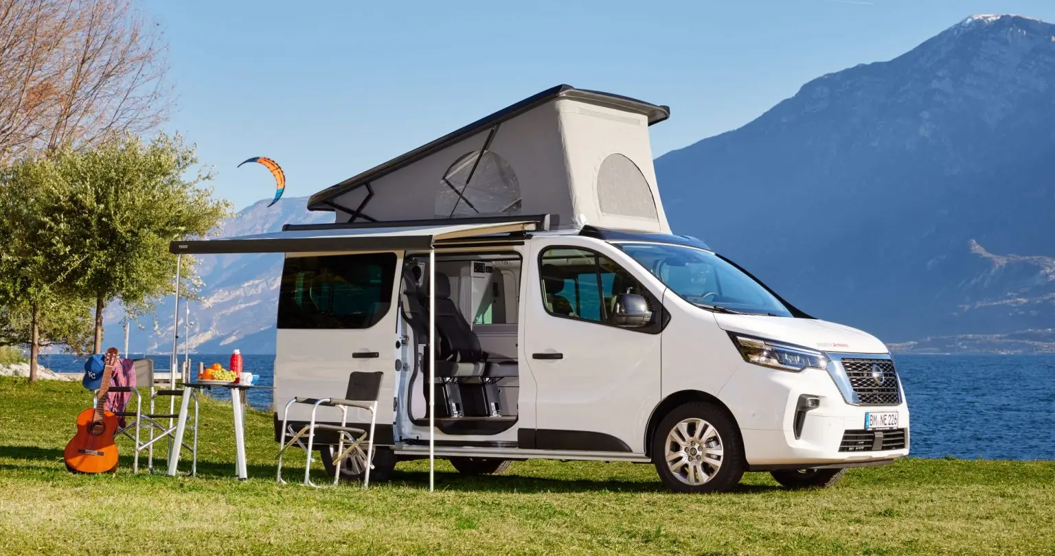 Weißer Nissan Primastar Campervan mit aufgeklapptem Aufstelldach und Campingmöbeln am See.
