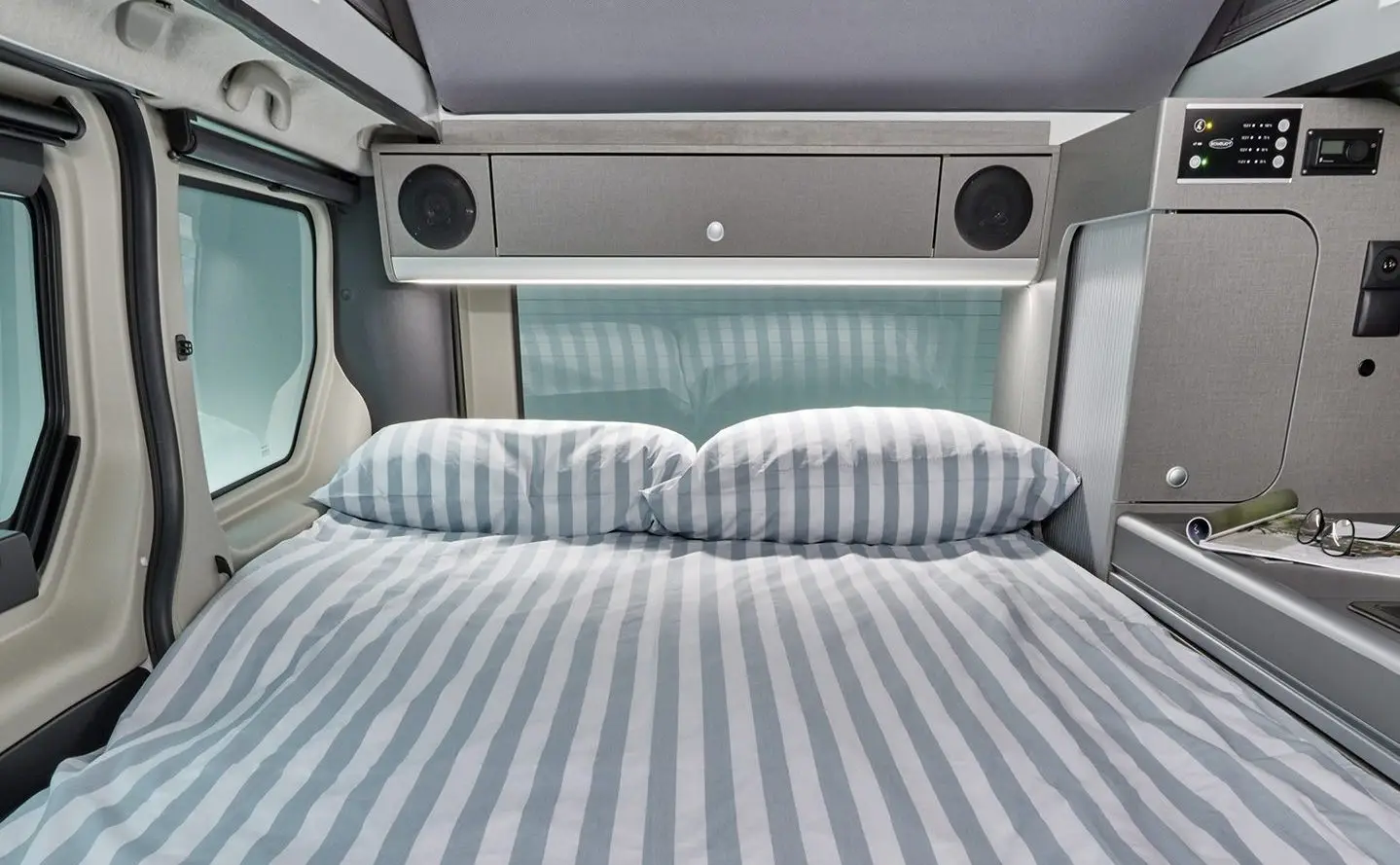 Bett im Heckbereich des Nissan Primastar Seaside mit gestreifter Bettwäsche und seitlichen Fenstern.