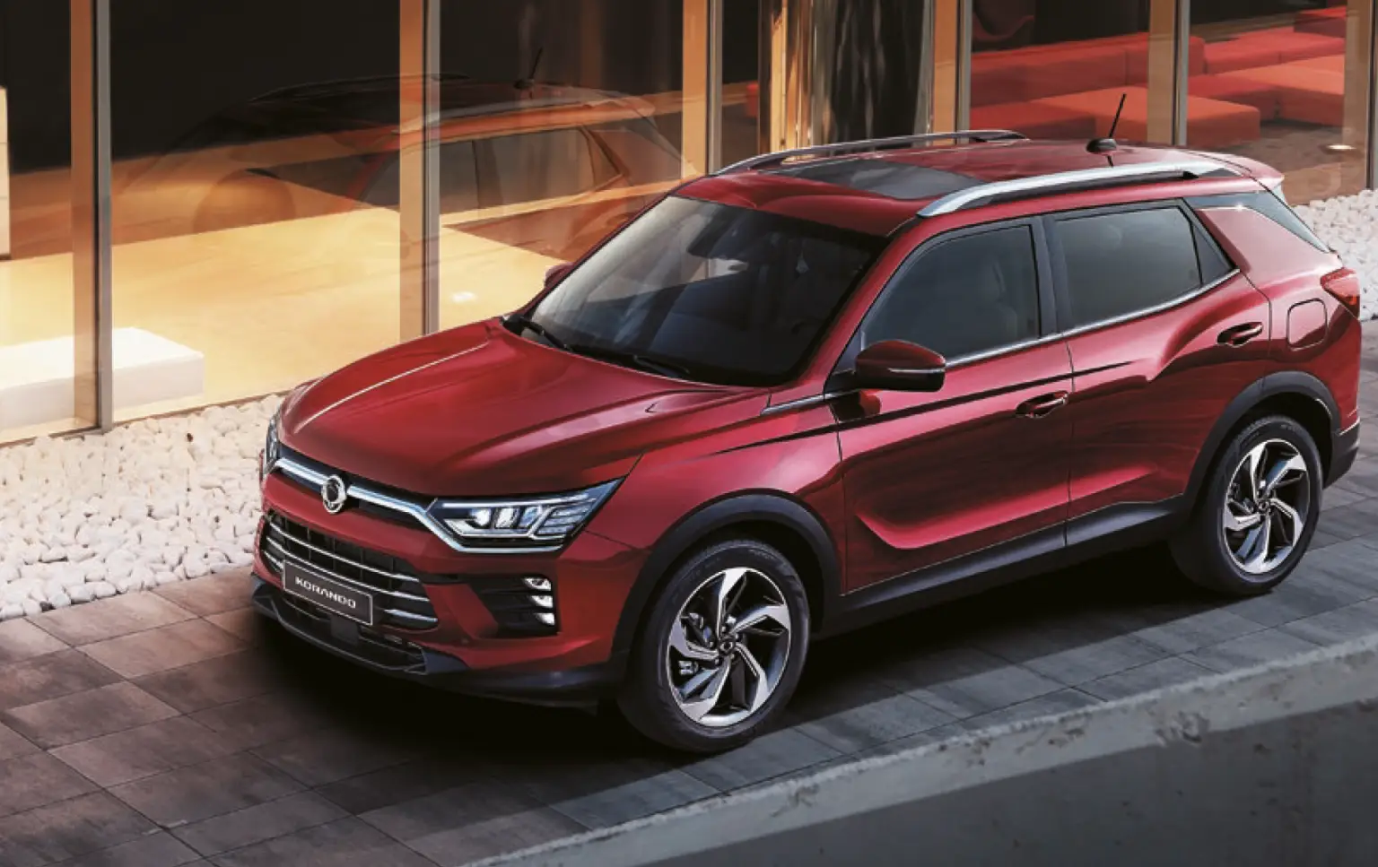 Roter KGM Korando SUV vor moderner Glasfassade bei Abendlicht.