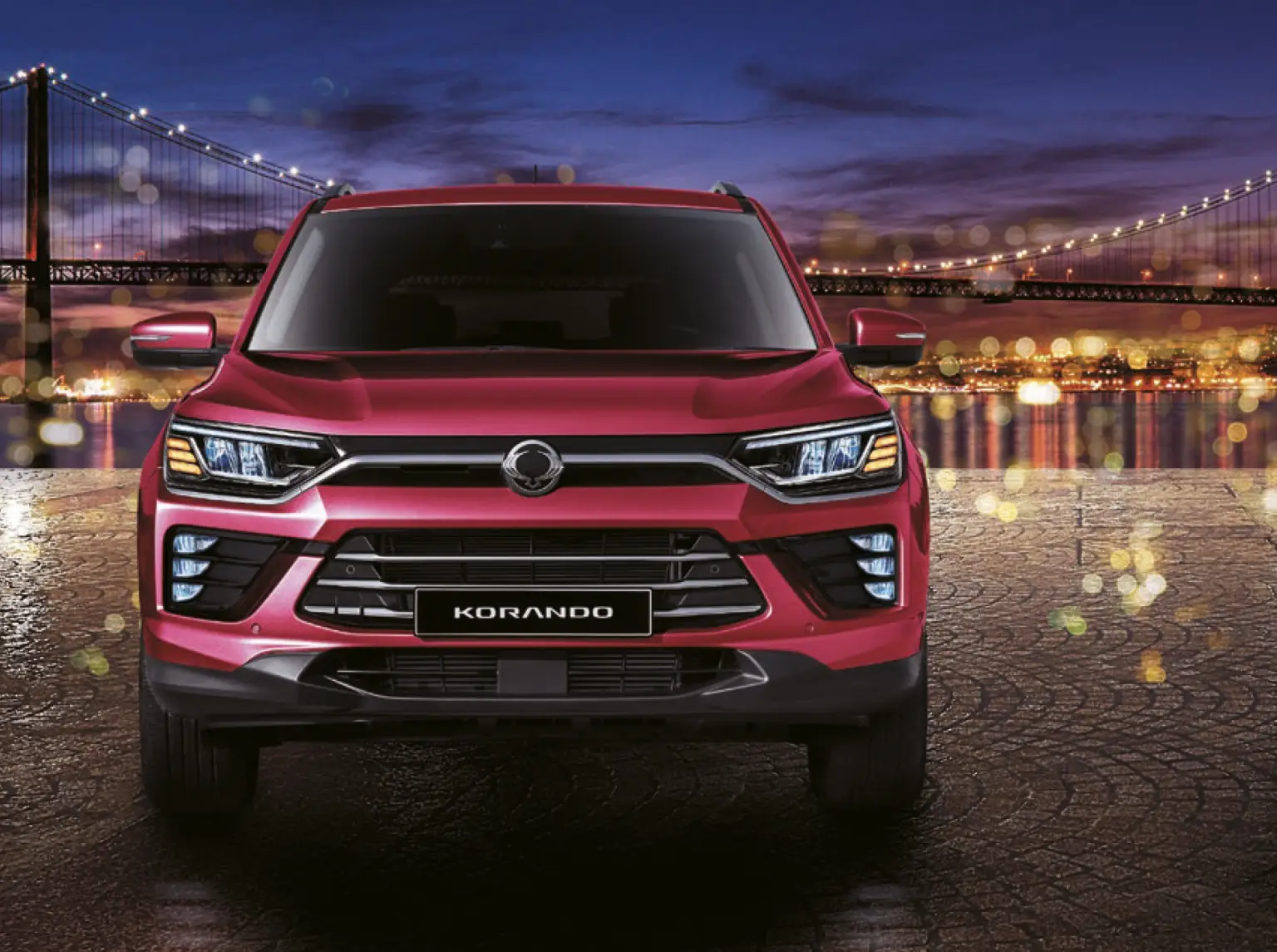 Roter KGM Korando SUV von vorne bei Nacht, im Hintergrund eine beleuchtete Hängebrücke und Stadtlichter.
