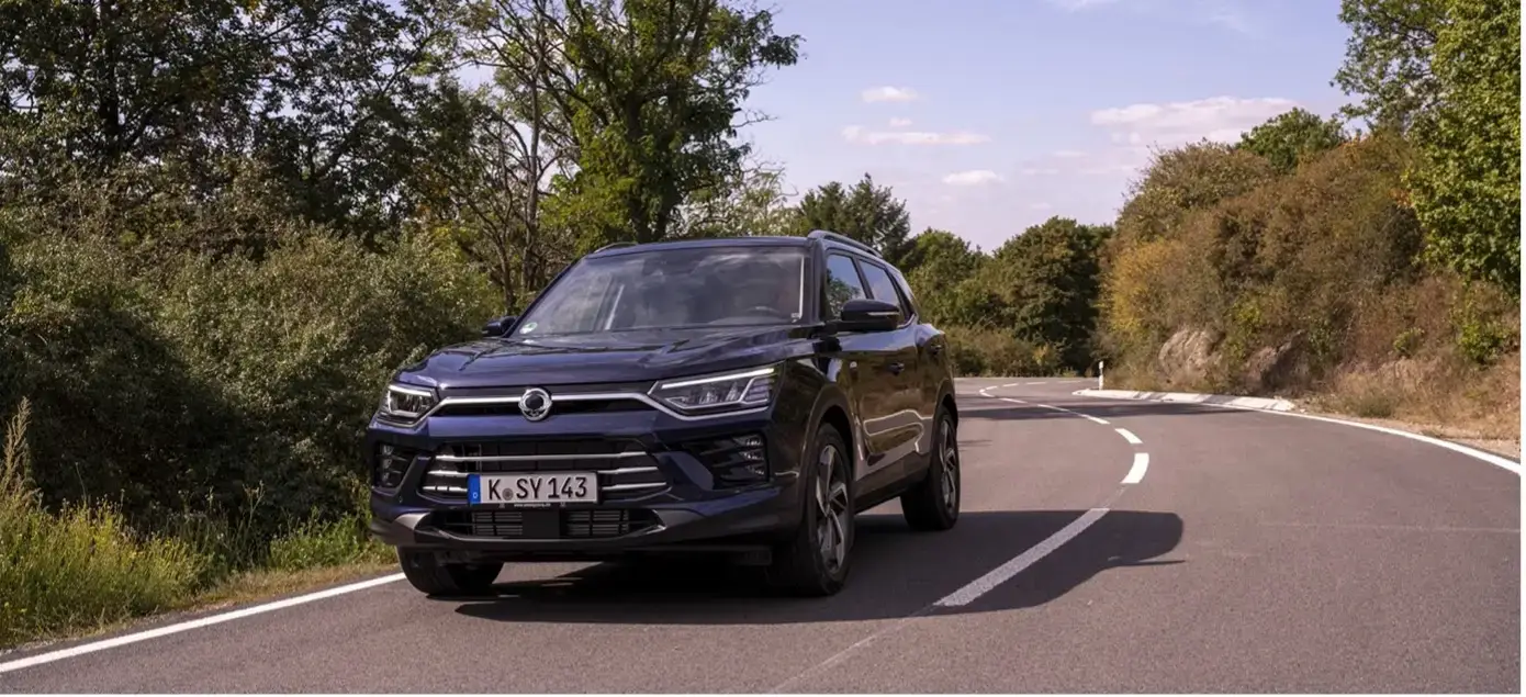 Ein KGM Korando in dunkler Lackierung fährt auf einer kurvigen Landstraße durch eine grüne Landschaft.
