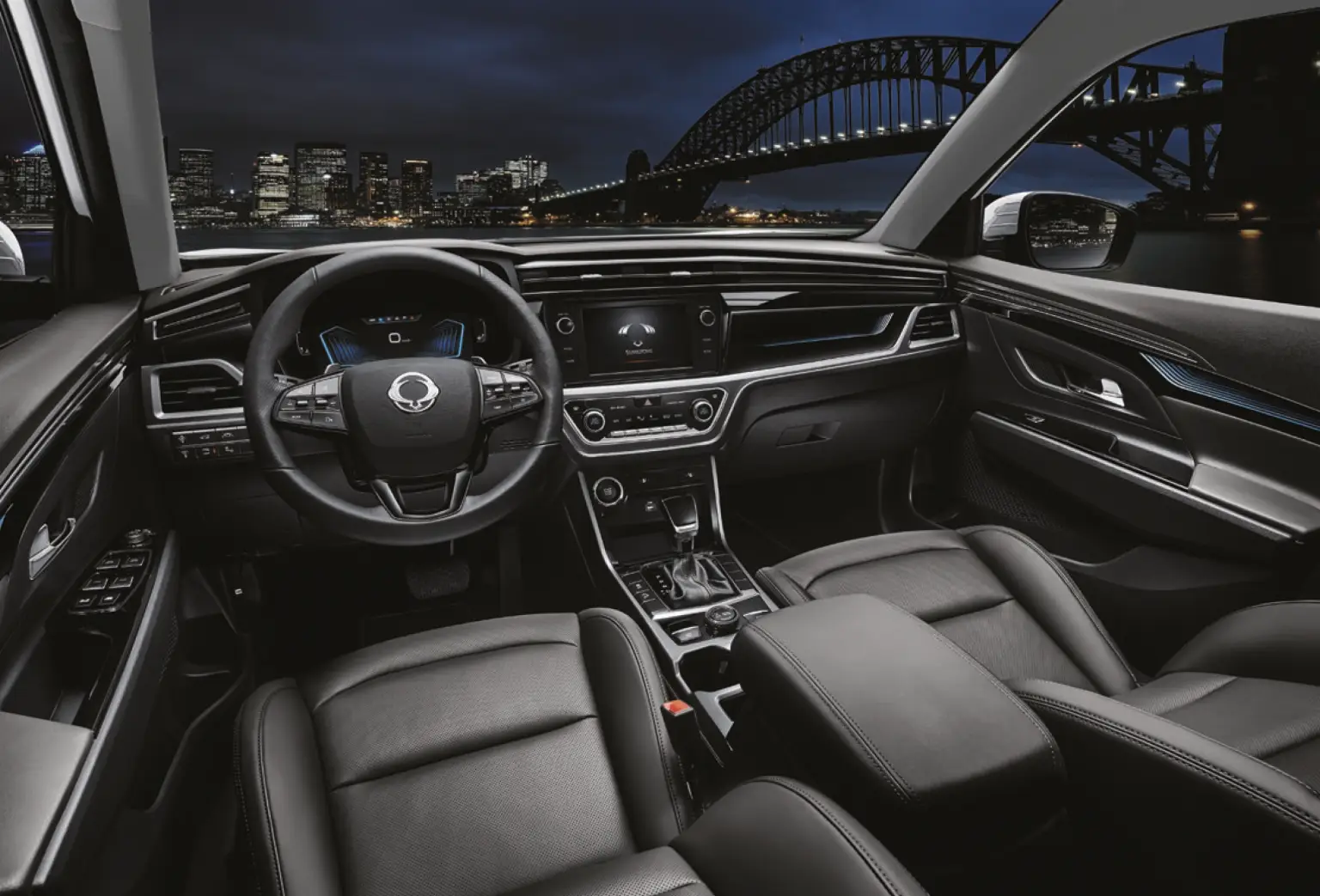 Modern gestalteter Innenraum des KGM Korando mit schwarzer Lederausstattung und digitalem Cockpit bei Nacht mit Blick auf eine Stadt und eine beleuchtete Brücke.