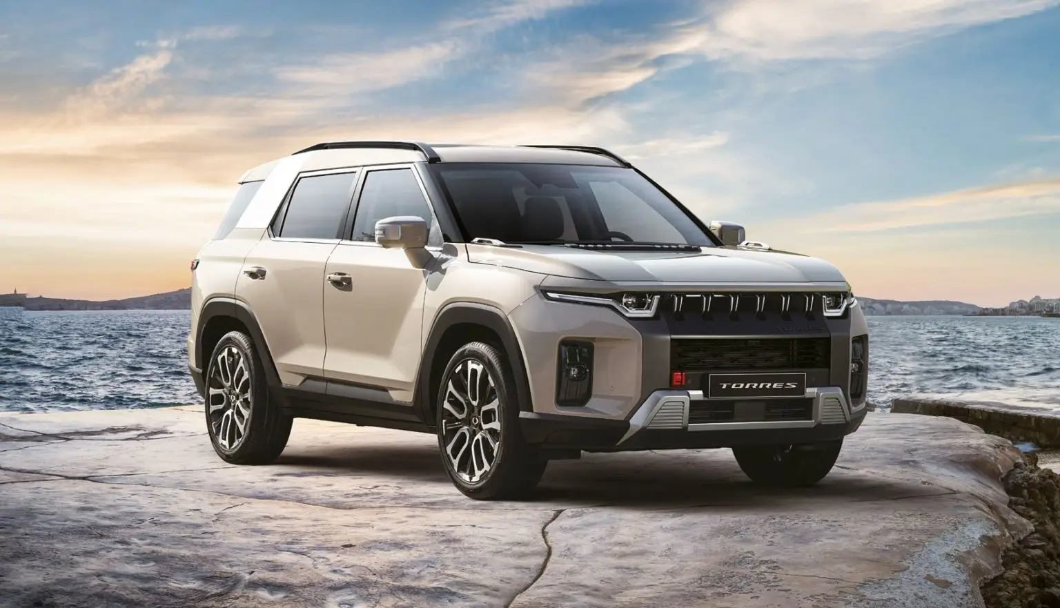 KGM Torres SUV in hellem Beige auf felsigem Untergrund vor Meereskulisse bei Sonnenuntergang.
