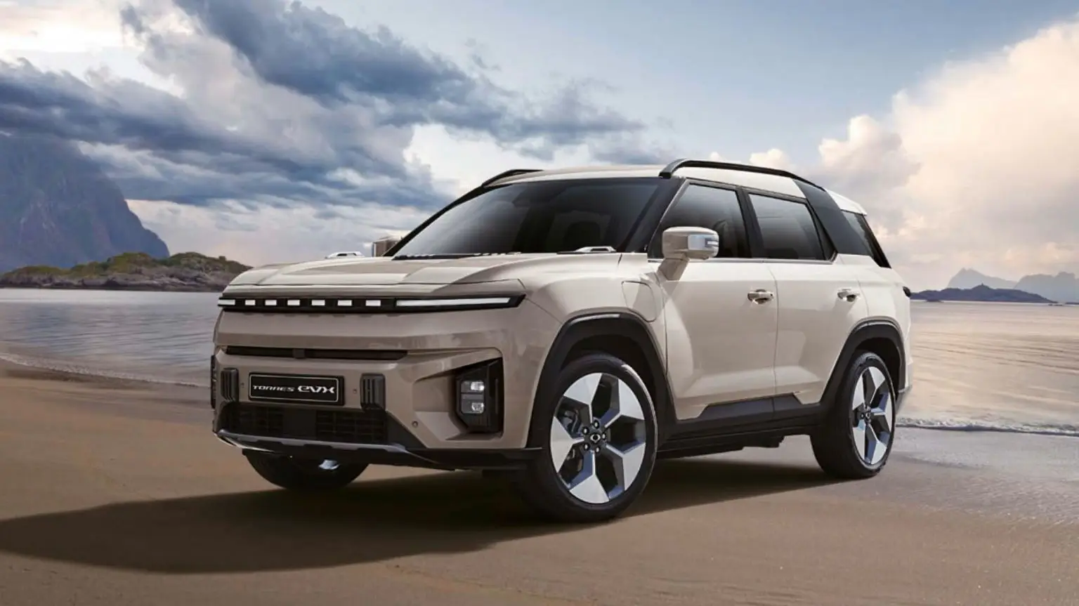 Der vollelektrische SUV KGM Torres EVX steht am Strand vor einer Küstenlandschaft mit Bergen und dramatisch bewölktem Himmel.